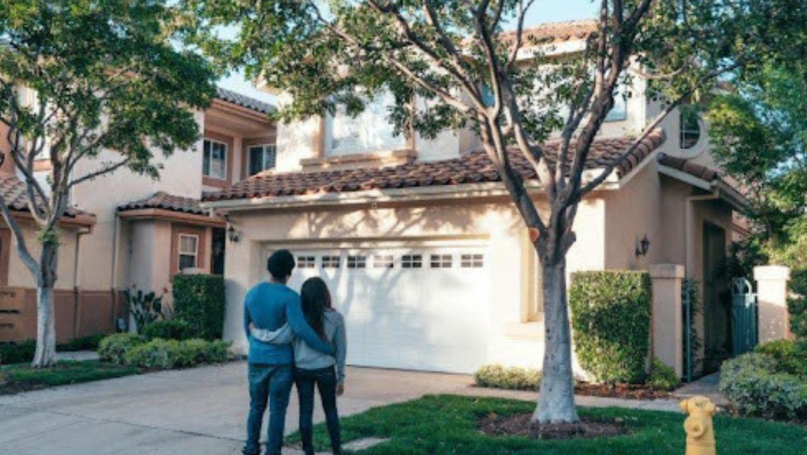 A couple stands arm in arm, facing a suburban house, symbolizing new opportunities and future plans in homeownership.