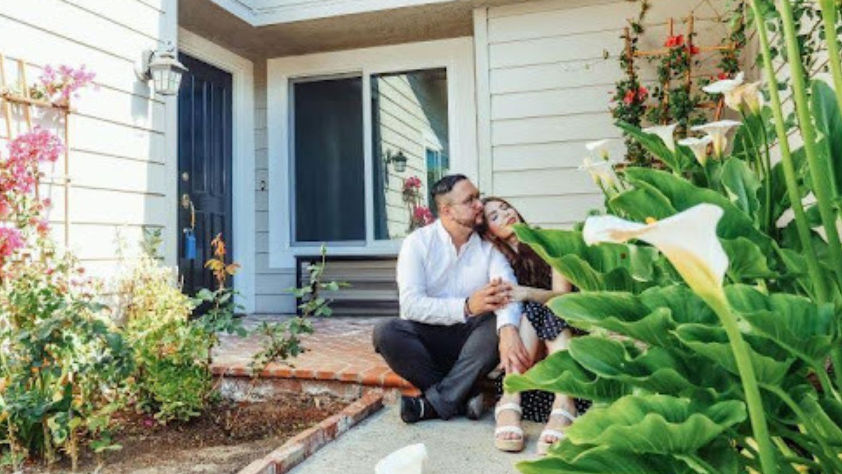 A smiling couple sits together by the entrance of a suburban house surrounded by flowers, symbolizing peaceful home life.