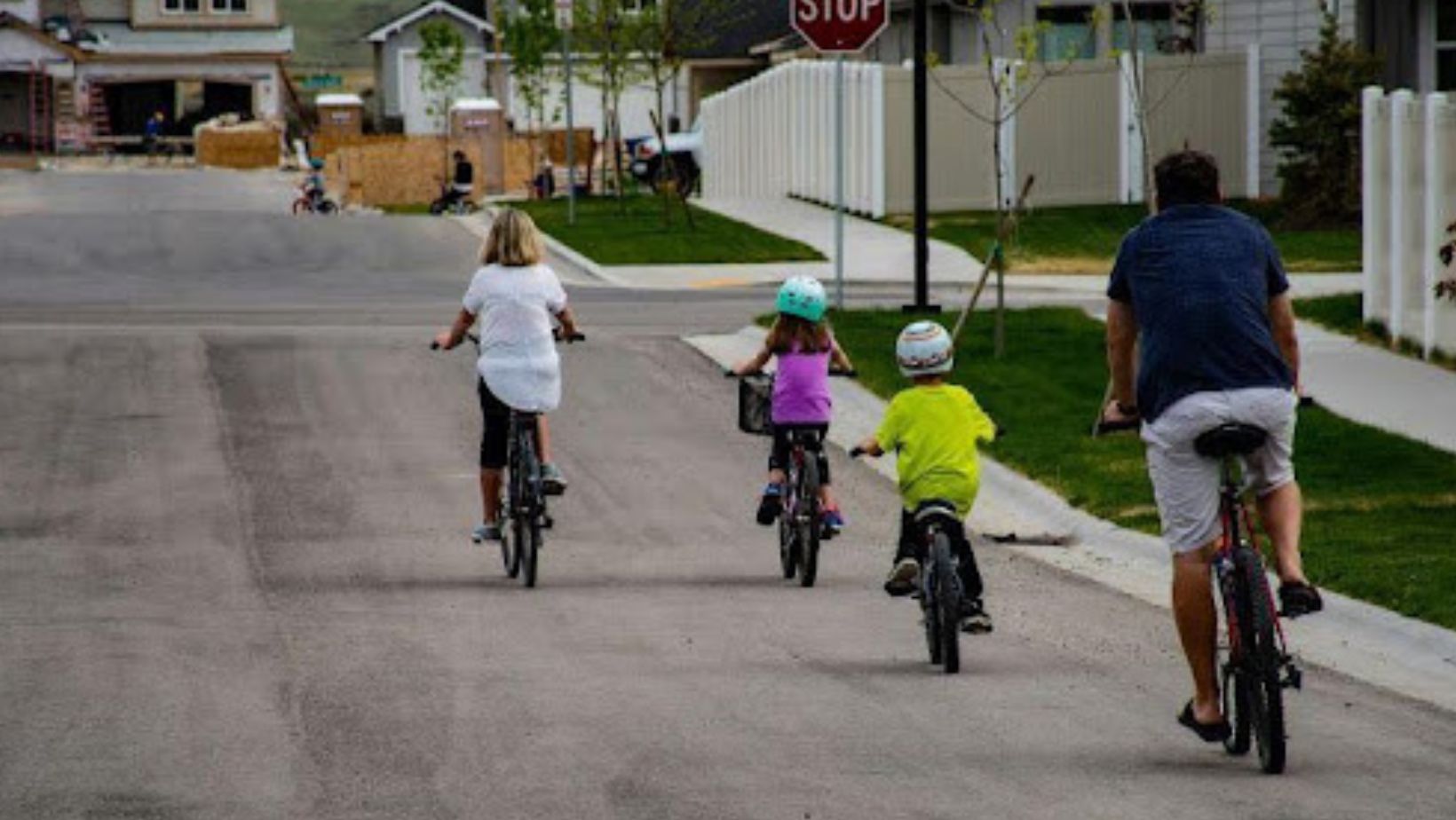 A family cycles down a suburban road with children wearing helmets, symbolizing active living and safe neighborhood spaces.